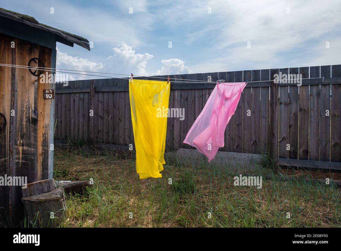 Saubere Kleidung wird an einem Seil getrocknet. Zwei Regenmäntel in rosa und gelben Farben flattern im Wind im Hof vor dem Hintergrund des Zauns. Stockfoto