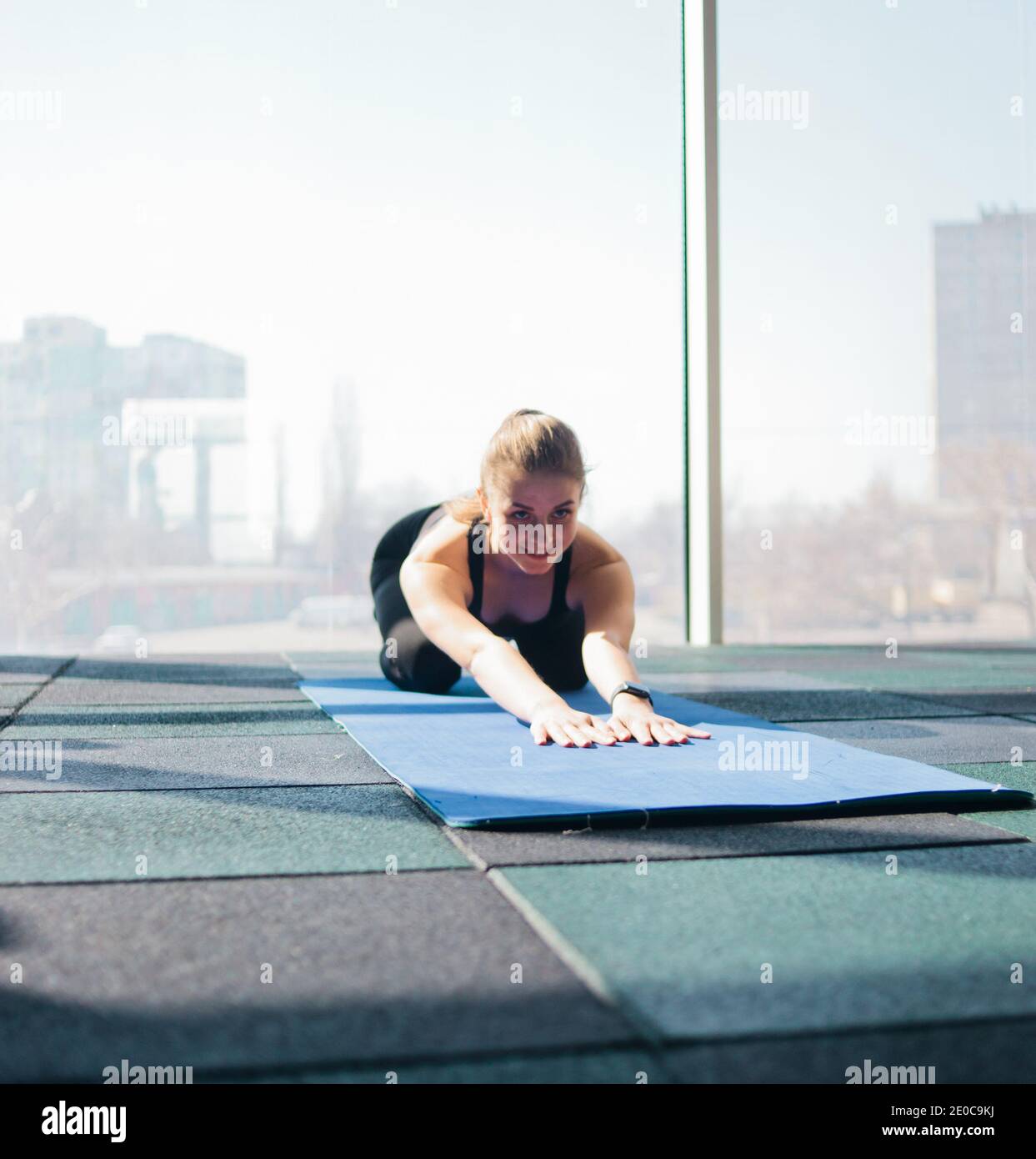 Junge Frau macht Stretching für den ganzen Körper auf Yoga-Matte vor dem Hintergrund der großen Panoramafenster in der Turnhalle. Yoga-Training Stockfoto
