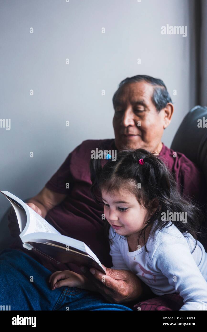 Ältere ethnische Großvater lesen interessante Geschichte im Buch für niedlich Kleines Mädchen beim Entspannen zusammen auf dem Sofa zu Hause Stockfoto