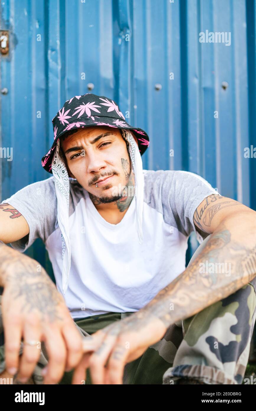 Junge tätowierte männliche Hipster in informeller Kleidung auf dem Boden sitzen In der Nähe von Metallzaun und Blick auf die Kamera Stockfoto