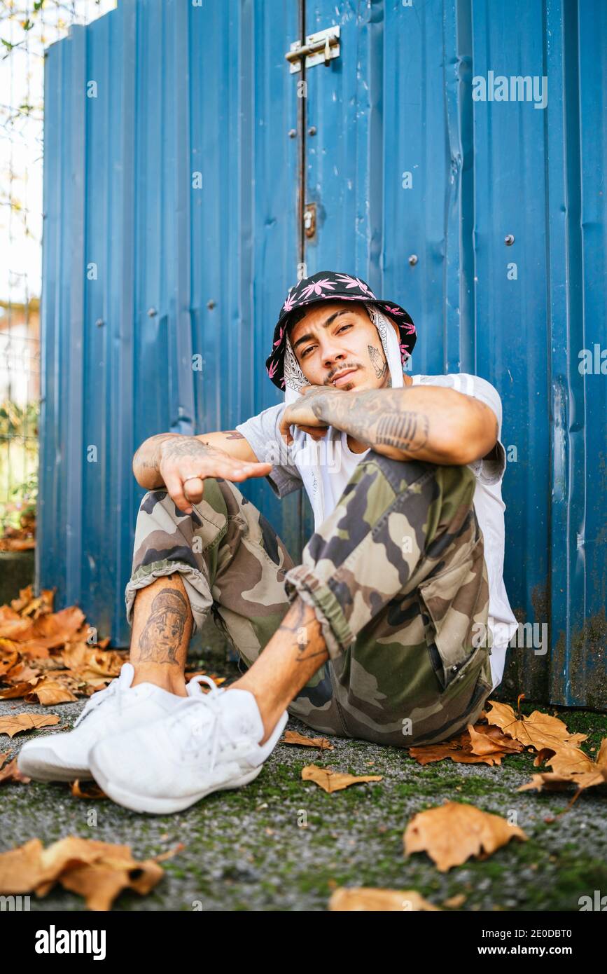 Ganzer Körper von jungen tätowierten männlichen Hipster in informeller Kleidung Sitzt auf dem Boden in der Nähe von Metallzaun und Blick auf die Kamera Stockfoto