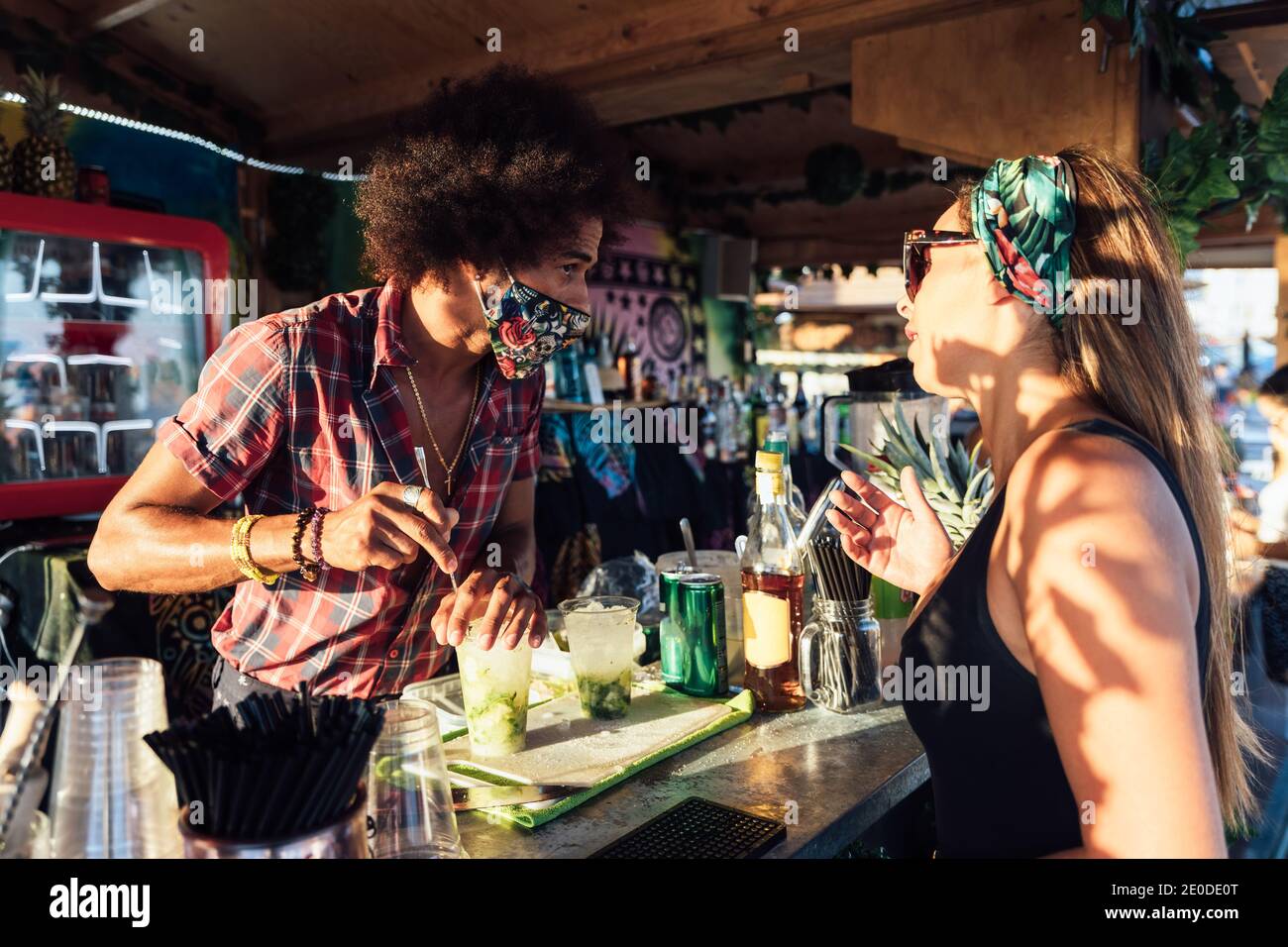 Afroamerikanischer Barkeeper macht erfrischenden alkoholischen Cocktail mit Eis und Lime für weibliche Gäste in der Bar im Freien am Sommerabend Stockfoto