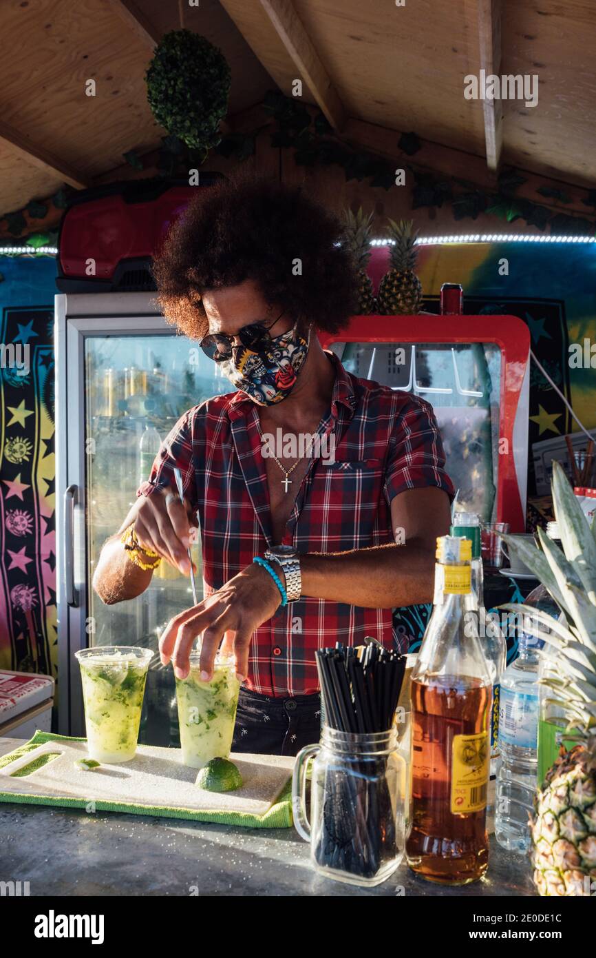 Afroamerikanischer Barkeeper in Sonnenbrille und Schutzmaske Mischung alkoholisch Trinken Sie mit Kalk und Eis, während Sie die Bestellung für den Kunden vorbereiten In der Bar im Freien Stockfoto