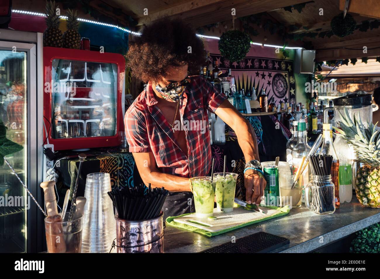 Afroamerikanischer Barkeeper in Sonnenbrille und Schutzmaske Mischung alkoholisch Trinken Sie mit Kalk und Eis, während Sie die Bestellung für den Kunden vorbereiten In der Bar im Freien Stockfoto