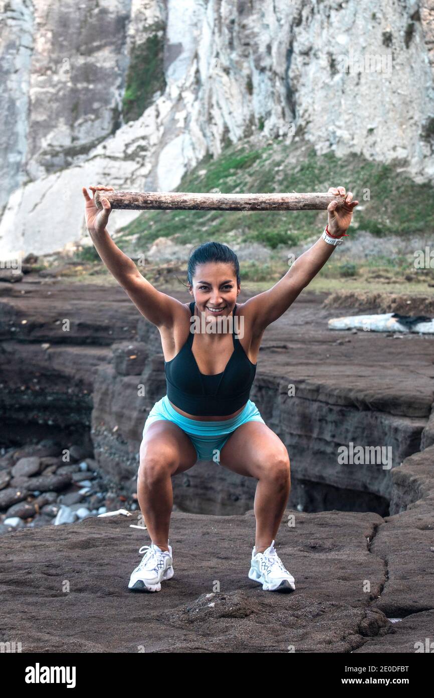 Entzückt athletische Frau in der Sportbekleidung hocken Übung mit Holz Halten Sie sich über den Kopf, während Sie am Meer trainieren und sich angucken Kamera Stockfoto