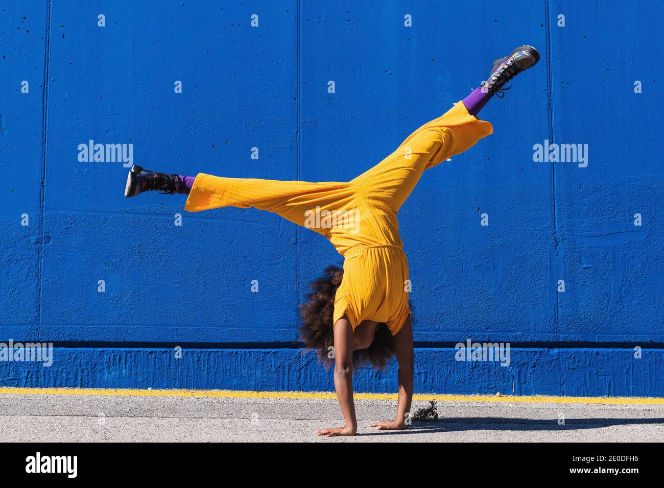 Ganzer Körper von afroamerikanischen Teenager-Mädchen in hellgelb Jumpsuit Handstand gegen blaue Wand auf der Straße Stockfoto