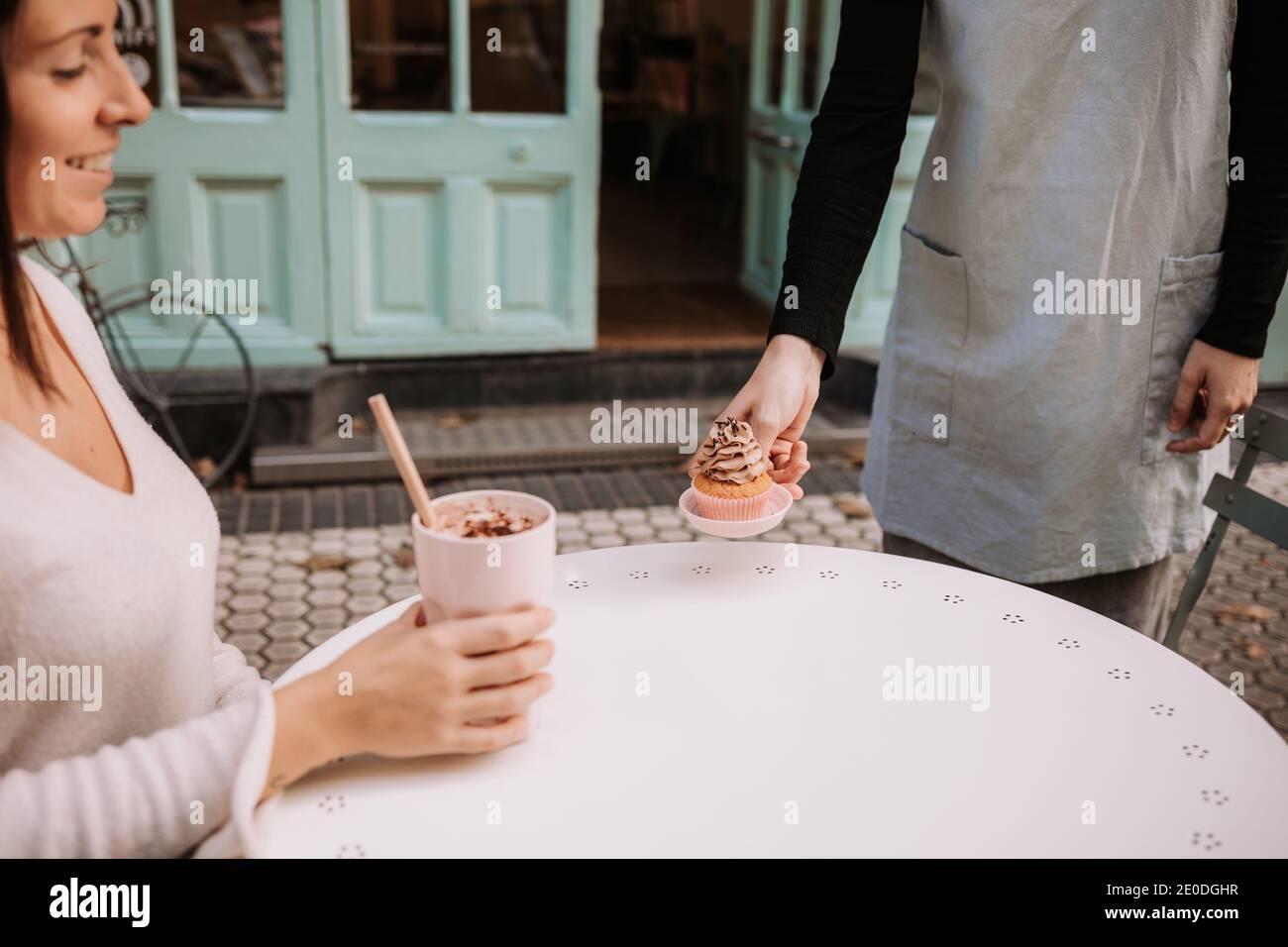Ernte Kellnerin Serving Cupcake zu glücklich weiblichen Kunden sitzen an Tisch mit Glas Frappe Kaffee in gemütlichen Süßwarencafe Stockfoto