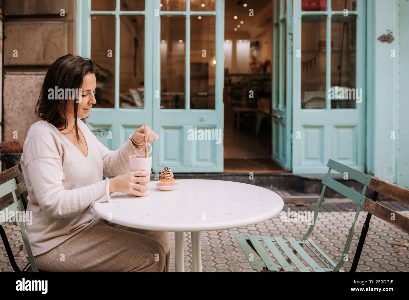 Seitenansicht der positiven erwachsenen Frau in legerer Kleidung sitzend Am Tisch auf der Terrasse der gemütlichen Cafeteria und genießen Frappe Kaffee und Cupcake Stockfoto