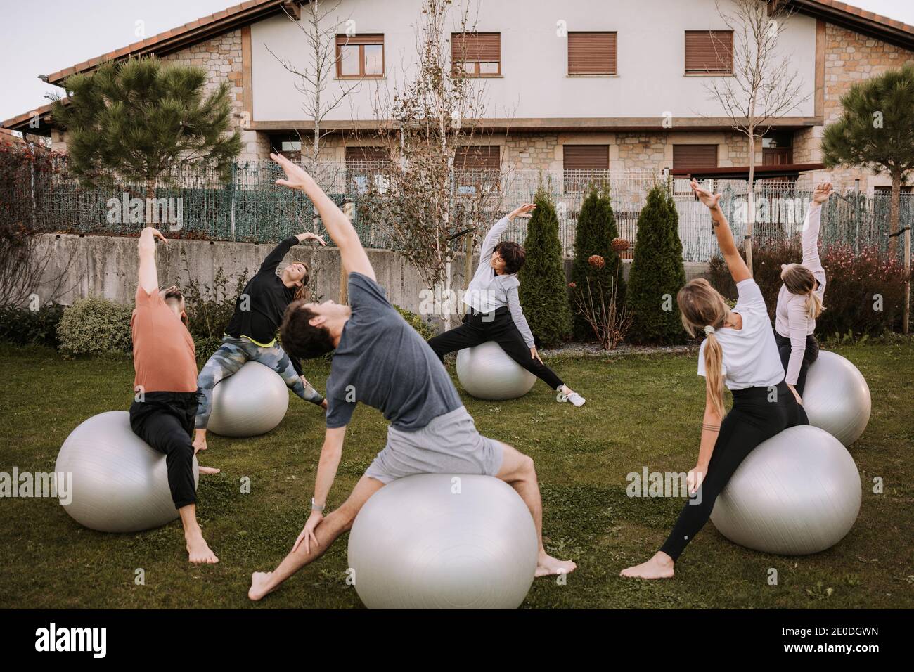 Gesellschaft von Menschen in Sportbekleidung sitzen auf fit Bälle auf Rasen und tun Seite biegen Übung mit angehobenem Arm während pilates-Schulung Stockfoto