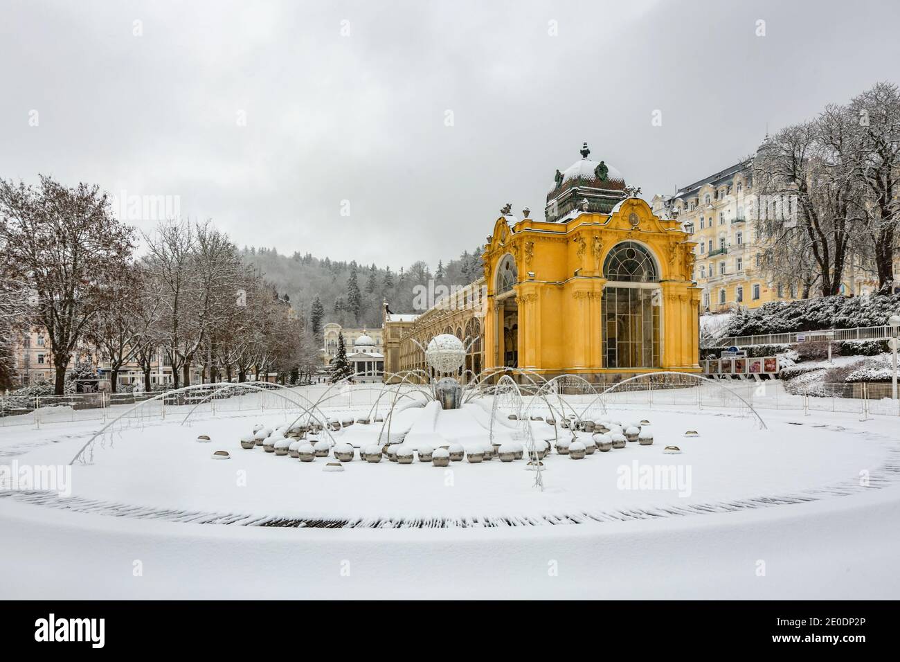 Marianske Lazne, Tschechische Republik - Dezember 29 2020: Der singende Brunnen im Vordergrund mit Schnee und gelb Maxim Gorki Kolonnade Haus bedeckt. Stockfoto