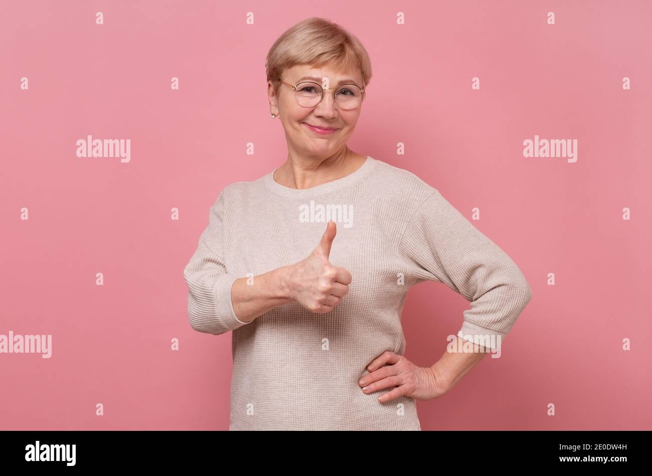Lächelnde ältere Frau zeigt Daumen nach oben. Studio auf rosa Wand aufgenommen Stockfoto