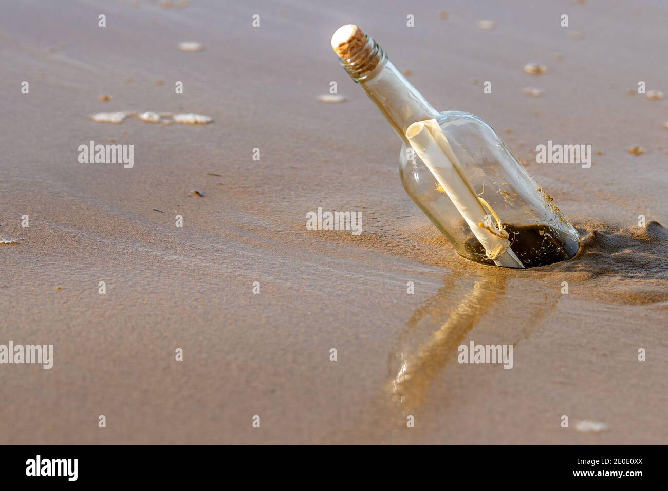 Eine Nachricht in einer korkierten Flasche, die an einem Strand aufgespült wurde. Die Flasche spiegelt sich im nassen Sand darunter wider. Die Flasche steht schräg. Sonniger Tag. Stockfoto