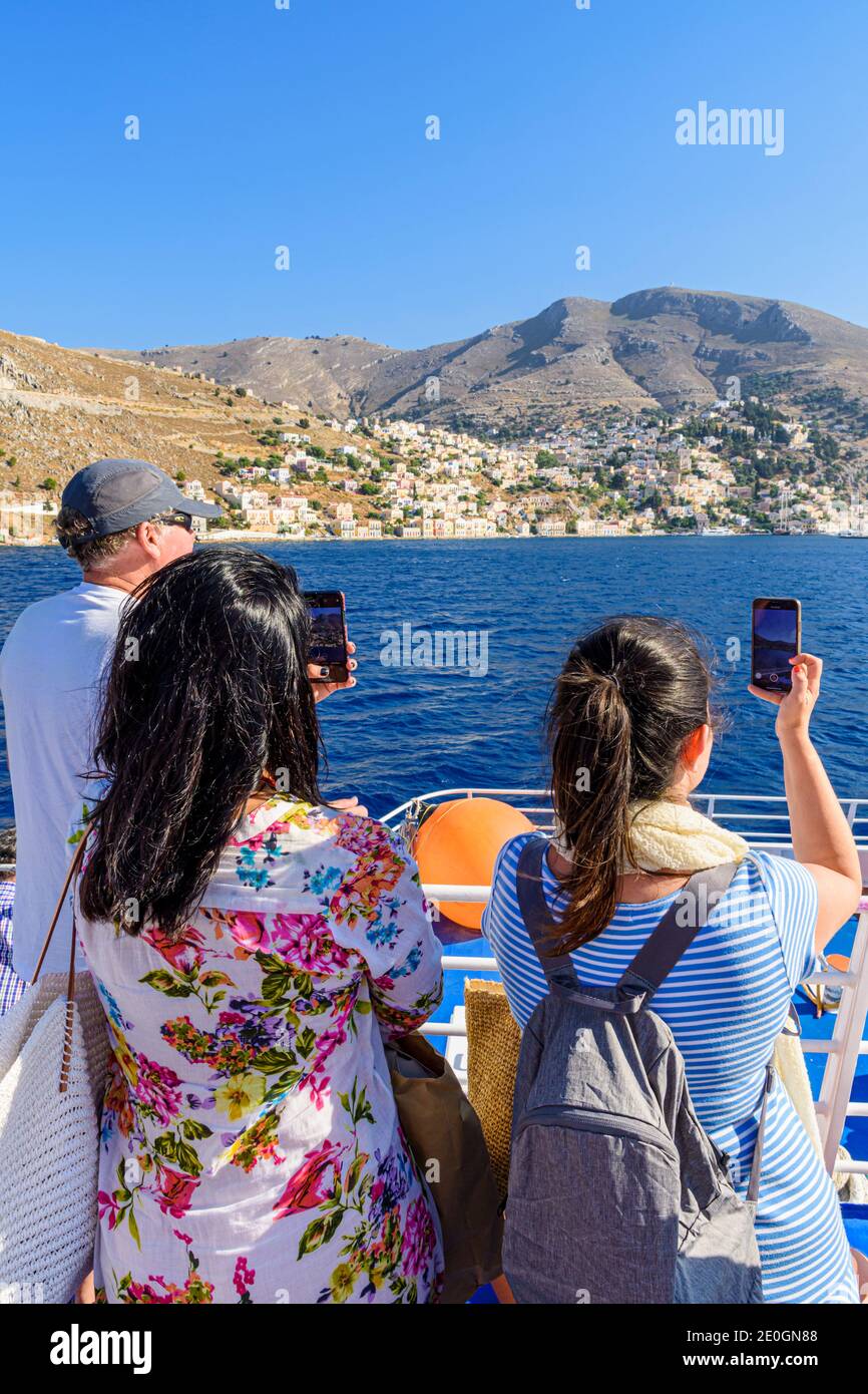 Reisende, die ihre Telefone benutzen, um den Blick vom Deck einer Fähre zu Filmen, wenn sie in der Hafenstadt Symi Island, Dodekanes, Griechenland ankommt Stockfoto
