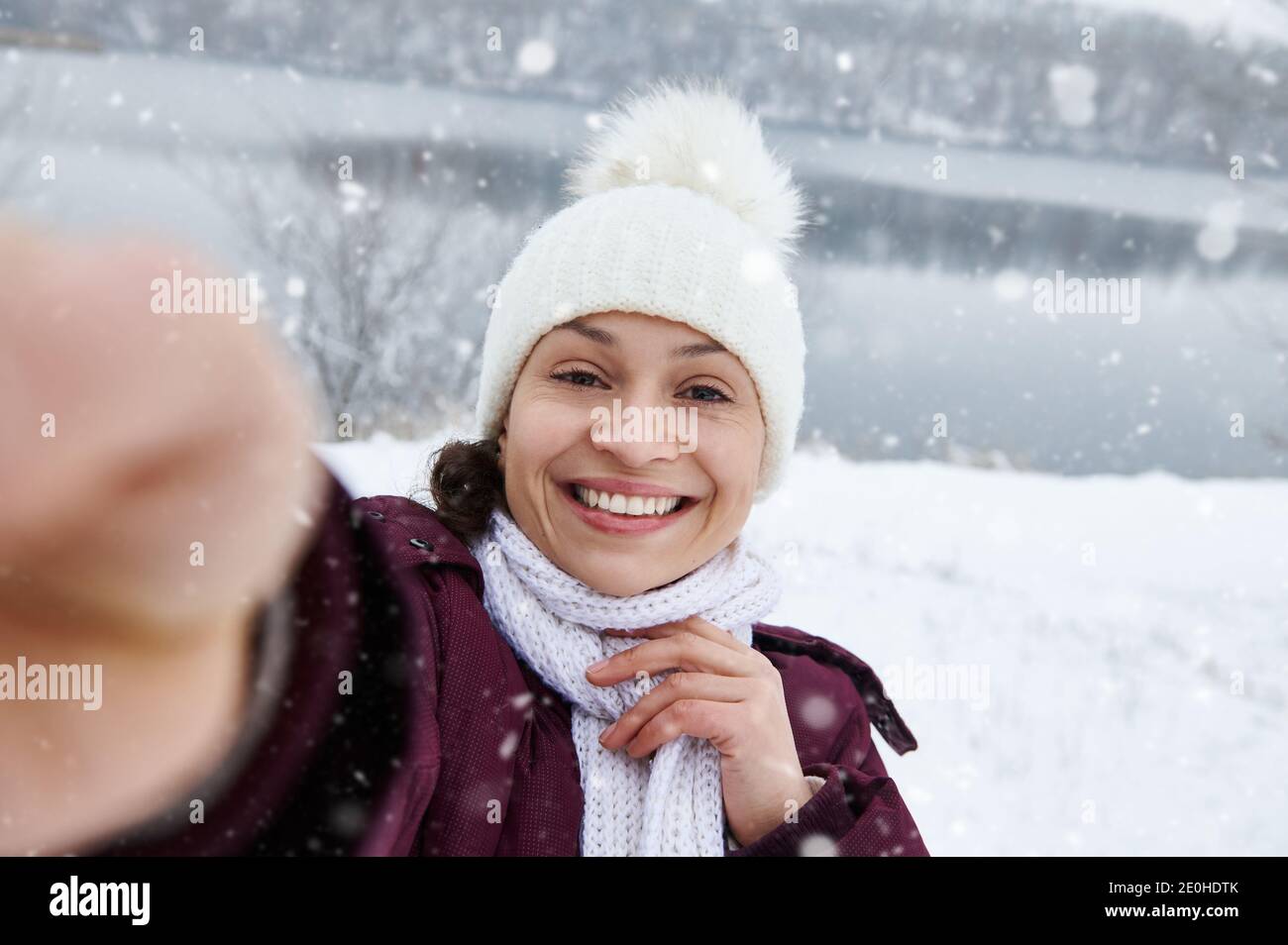 Nahaufnahme einer fröhlichen Brünette Frau lächelt an der Kamera, während Machen Sie ein Selfie, genießen Sie einen fallenden Schnee auf dem Hintergrund Von schneebedeckter Landschaft Stockfoto