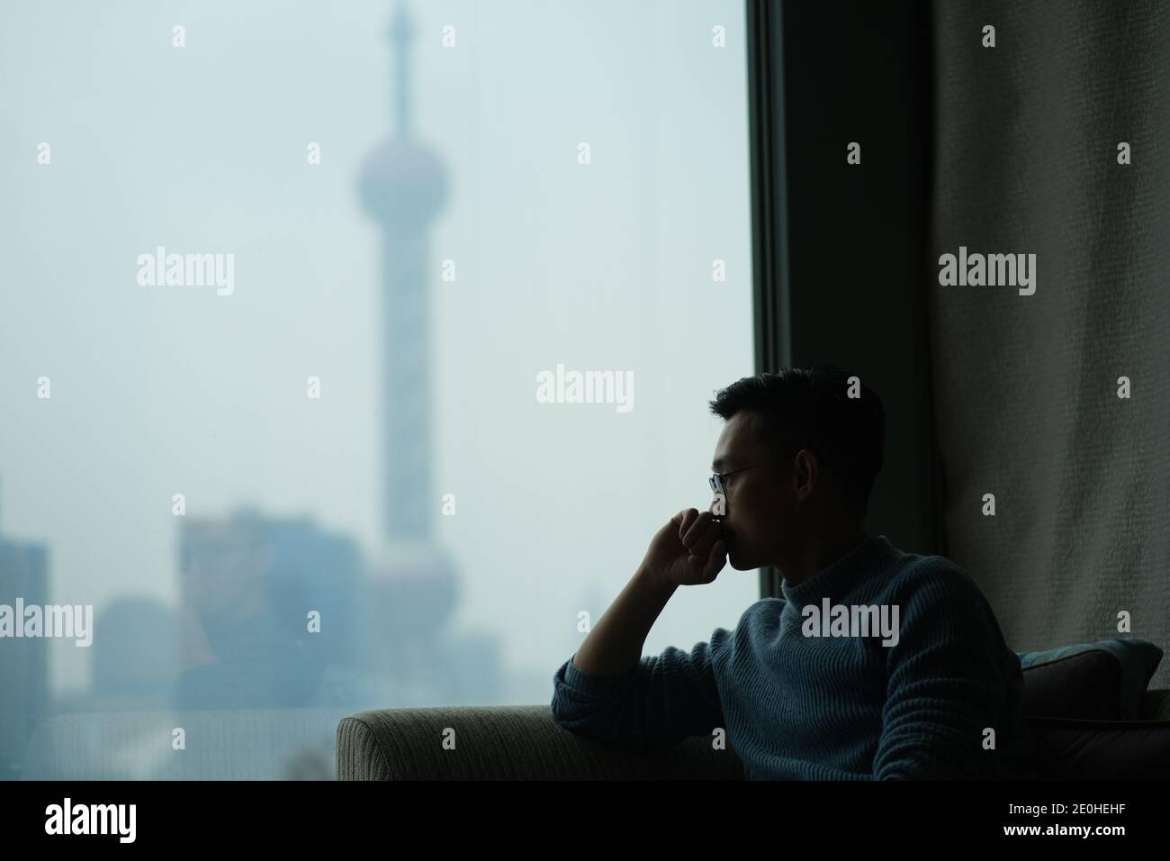 Mitten in der Aufnahme eines asiatischen jungen Mannes, der nachdenklich neben dem Fenster sitzt, verwischen Sie ein modernes Wahrzeichen aus dem Fenster Stockfoto