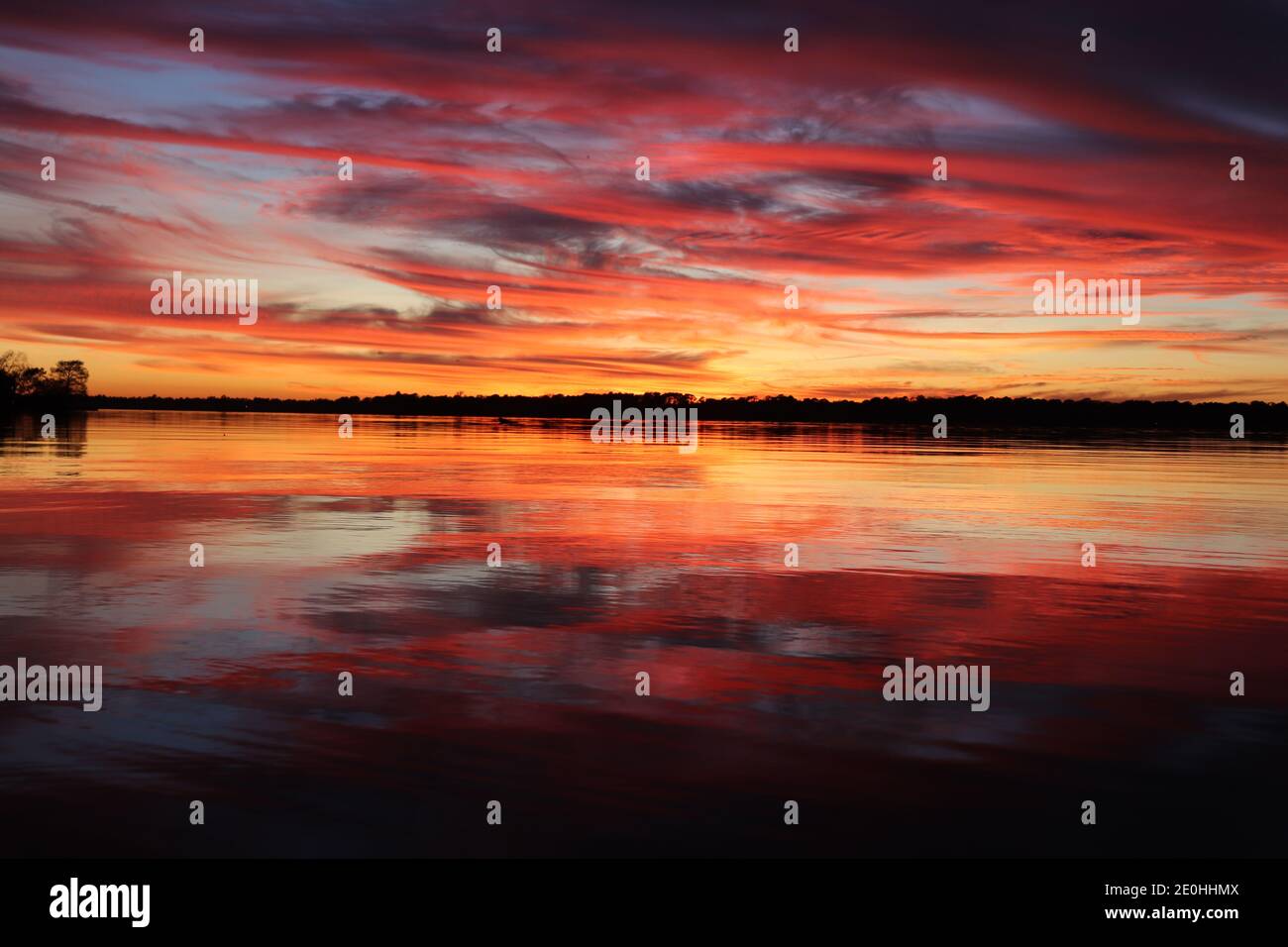 Sonnenuntergang mit Spiegelreflexen auf der Wasseroberfläche. Lebendige Farben von Rot und Gold wirbeln vom blauen Himmel bis zum ruhigen baumgesäumten Horizont. Stockfoto