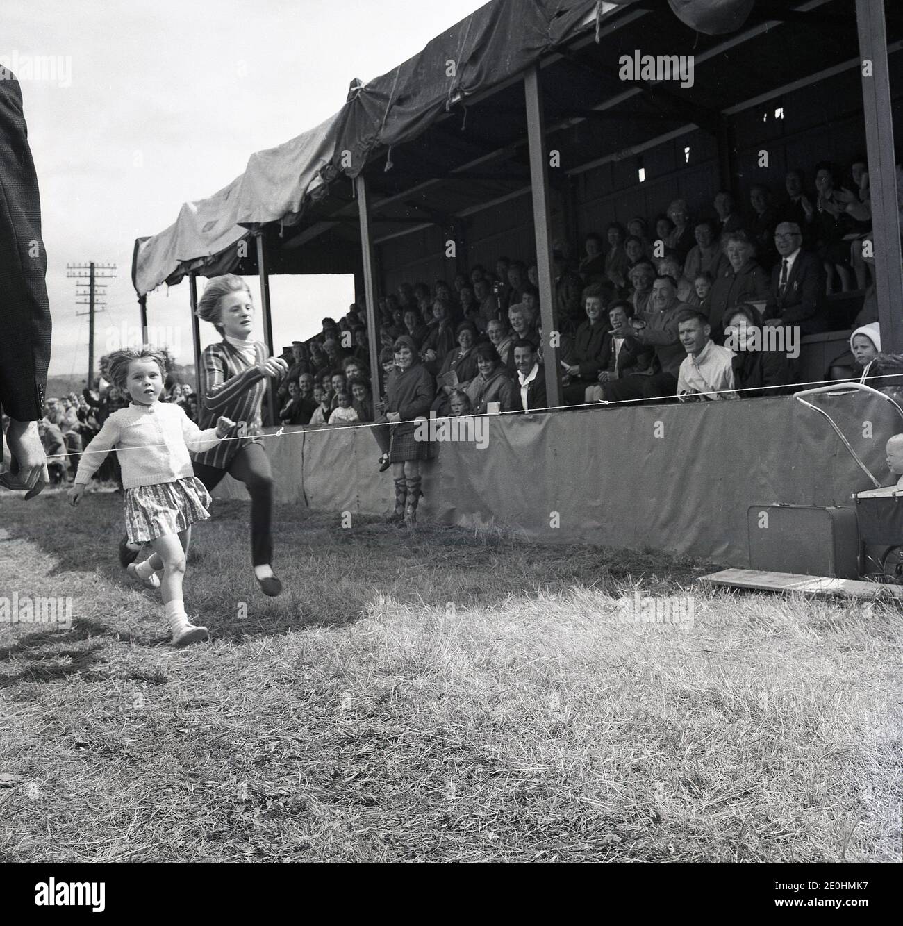 Zwei kleine Kinder, die in den 1960er Jahren bei einem lustigen Rennen auf dem Rasen Rennen, beobachtet von einer Menge von Schaustellern, die auf einer geschützten hölzernen Tribüne bei den Highland Games, Highlands, Schottland, Großbritannien, sitzen. Stockfoto