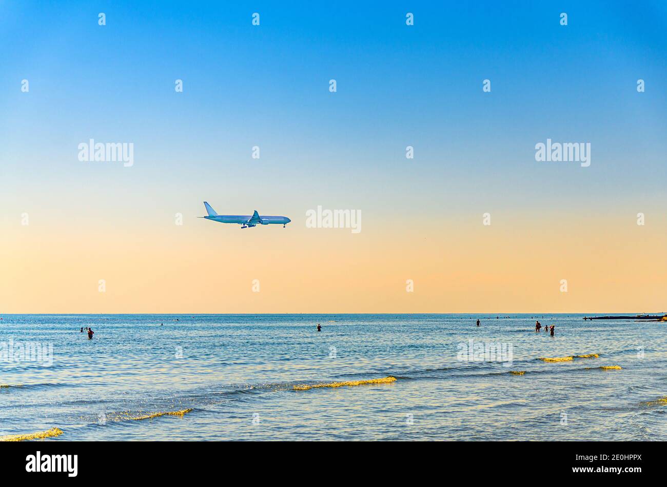 Flugzeug, das tief über dem Meer fliegt, und Menschen, die im Wasser schwimmen, klarer, blau-oranger Himmel bei Sonnenuntergang, Flugzeug, das sich darauf vorbereitet, am Flughafen Larnaca oben zu landen Stockfoto