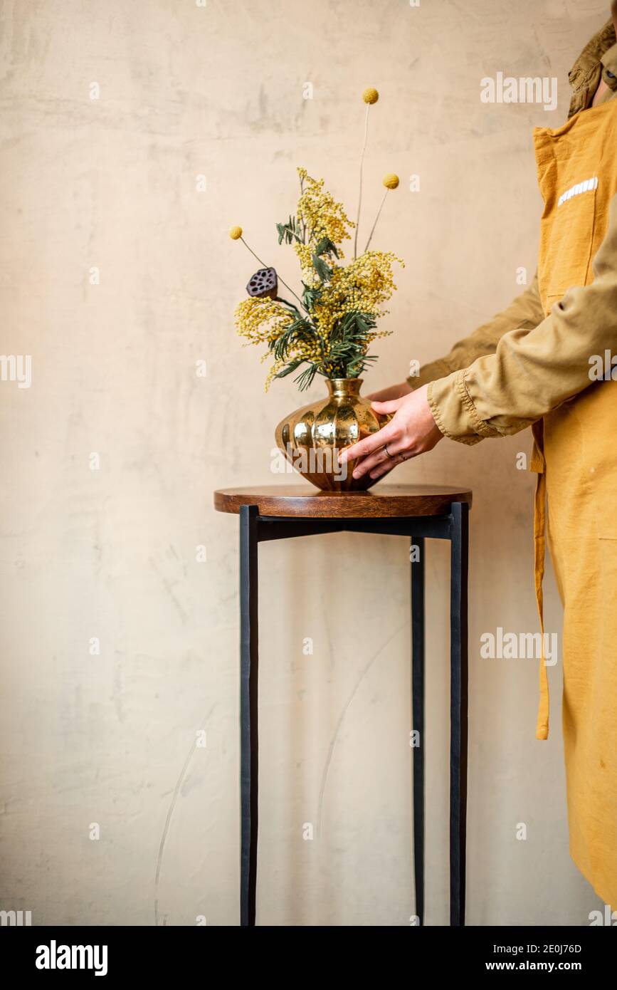Weibliche Dekokartierung nach Hause mit Komposition aus frischen Blumen und Kräutern in einer goldenen Vase auf einem beigen Wandhintergrund. Bouquet von Acacia, Dealbata, Mimosa, nelumbo und Craspedia Stockfoto
