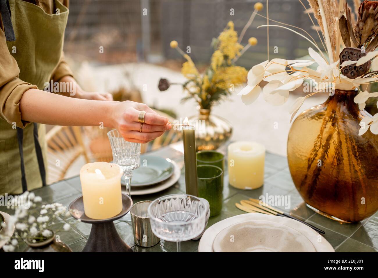 Junge Frau zündet Kerzen an, während sie den Esstisch in Natur dekorieren Boho-Stil in Grüntönen im Freien Stockfoto