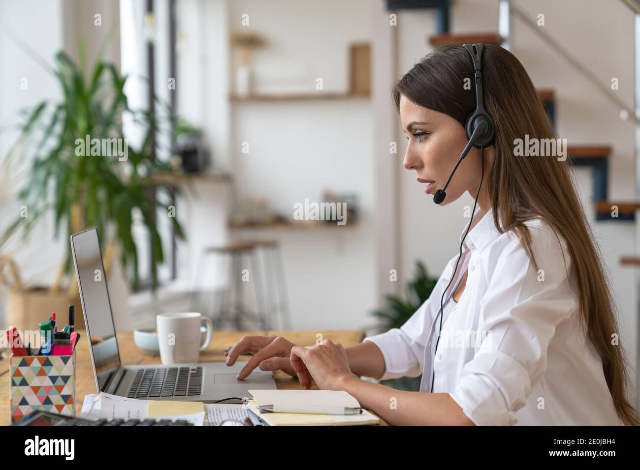 Seitenansicht des fokussierten Anrufers oder des Telefonanbieters an der Rezeption in Ohrhörern, die am Tisch sitzen, im Videochat sprechen, von zu Hause aus arbeiten. Di Stockfoto