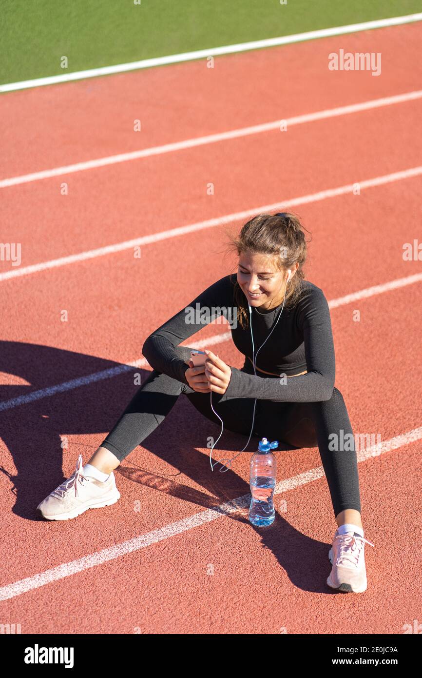 Fit Frau in Sportbekleidung Ruhe nach dem Training oder Laufen, sitzen auf einem Laufband Gummiband, mit und hört Musik mit kabelgebundenen Kopfhörer auf sma Stockfoto
