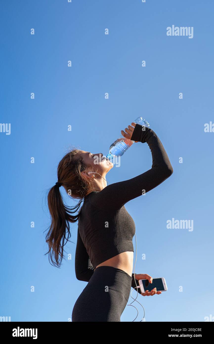 Durstige junge, fitstige Frau trinkt Wasser aus der Plastikflasche, ruht sich nach dem Joggen aus, hält Handy in der Hand, hört Musik. Blauer Himmel w Stockfoto