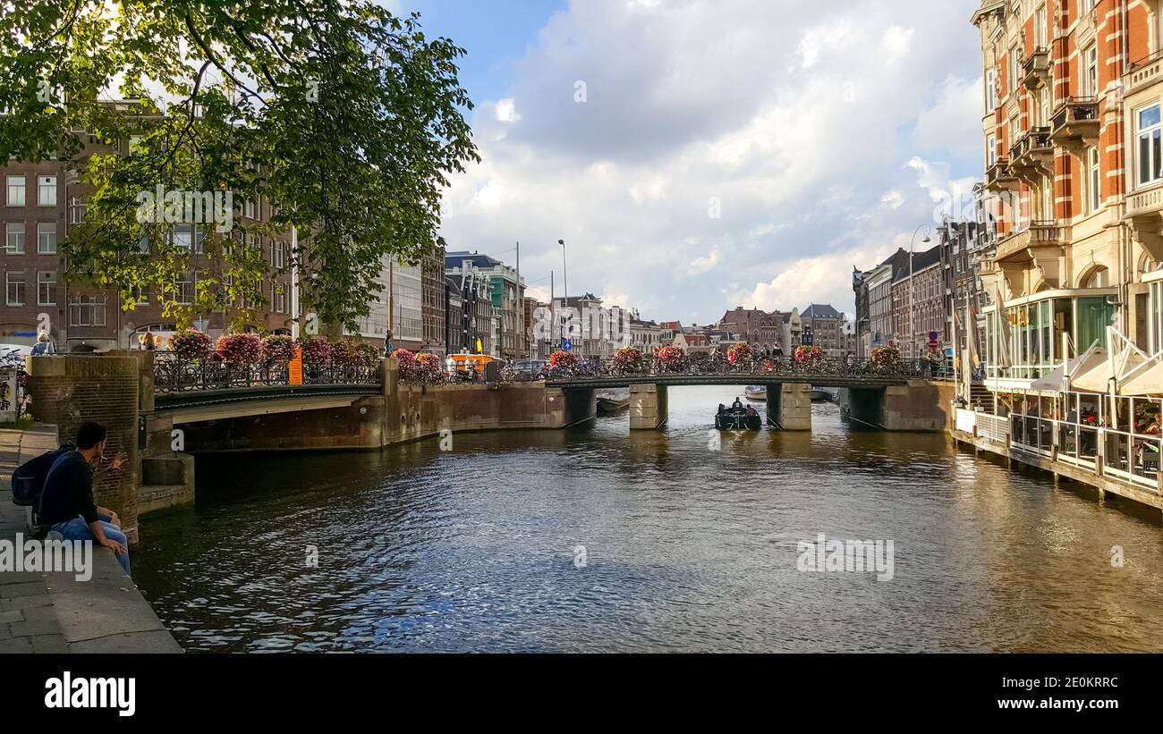 Malerische Lage an einem Kanal in Amsterdam mit einem Waterfront Cafe, Brücke mit Fahrrädern und Blumen, ein Boot und ein junges Paar genießt die Aussicht Stockfoto