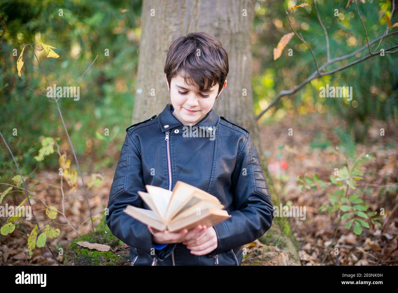 11-jähriger Junge liest ein Buch, das sich im Wald an einen Baum lehnt - ein Buch im Freien lesen - Kinder erziehen, um zu lesen. Stockfoto