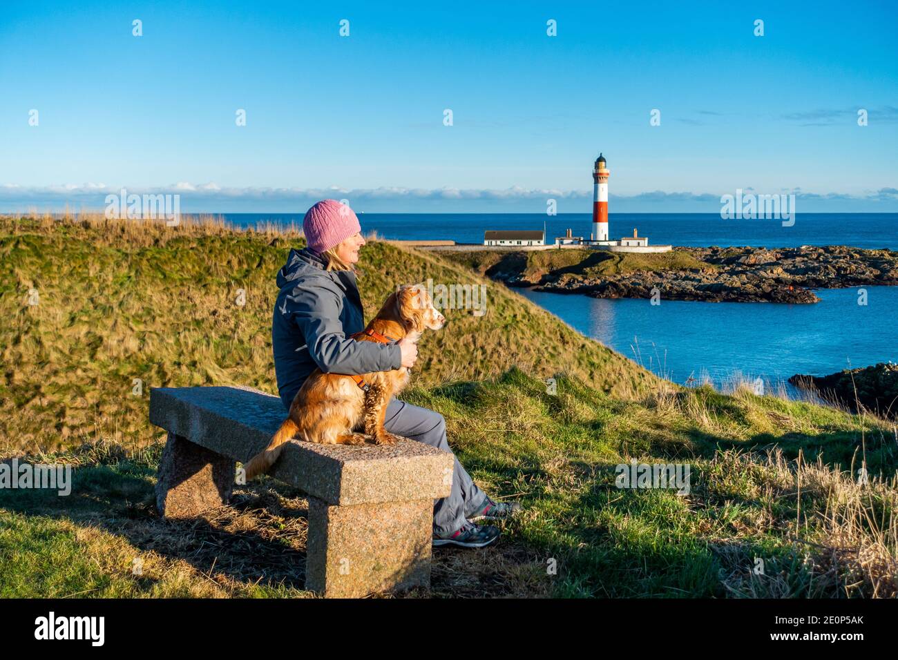 Ein Hundespaziergänger ruht sich auf einer Bank aus, die den rot-weißen Leuchtturm im Dorf Boddam bei Peterhead in Aberdeenshire, Schottland, Großbritannien, überblickt Stockfoto