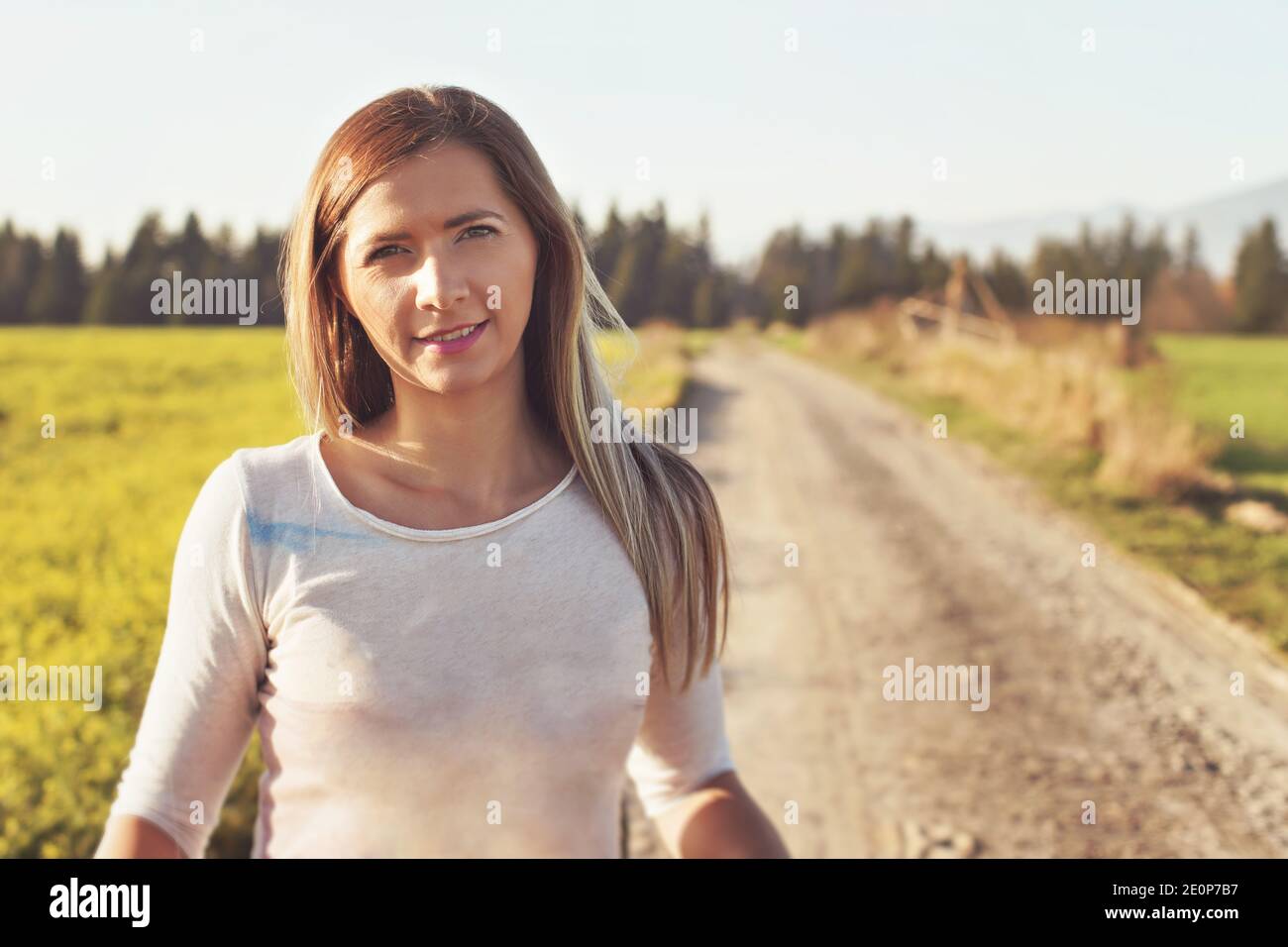 Porträt einer jungen Frau auf staubigen Landstraße, beleuchtet von der Nachmittagssonne, verschwommene Felder und Wald im Hintergrund, Foto getönten warmen Farben Stockfoto