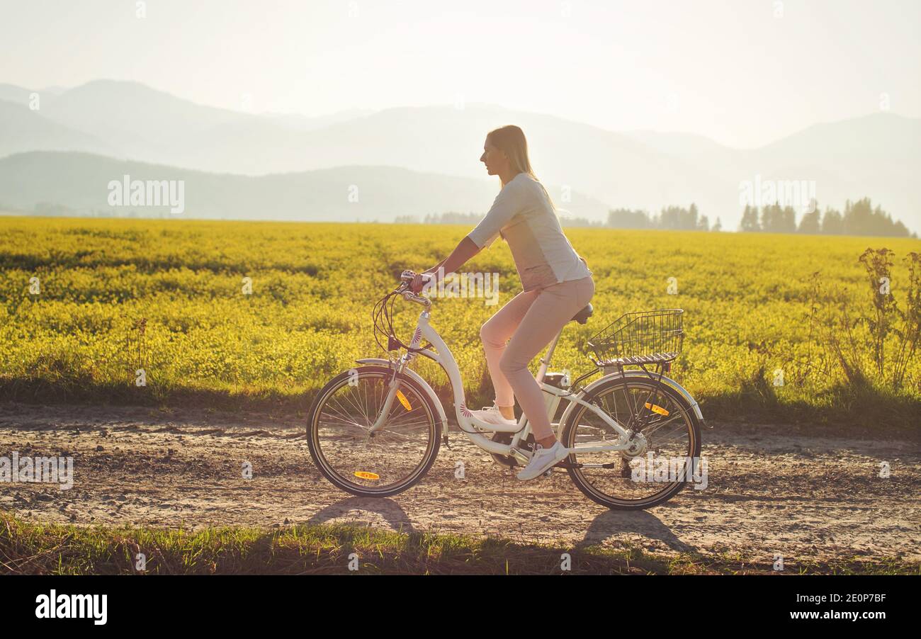 Junge Frau fährt Elektro-Fahrrad über staubige Landstraße, starke Nachmittag Sonne Hintergrundbeleuchtung im Hintergrund leuchtet auf gelben Blumen Feld, Blick von der Seite Stockfoto