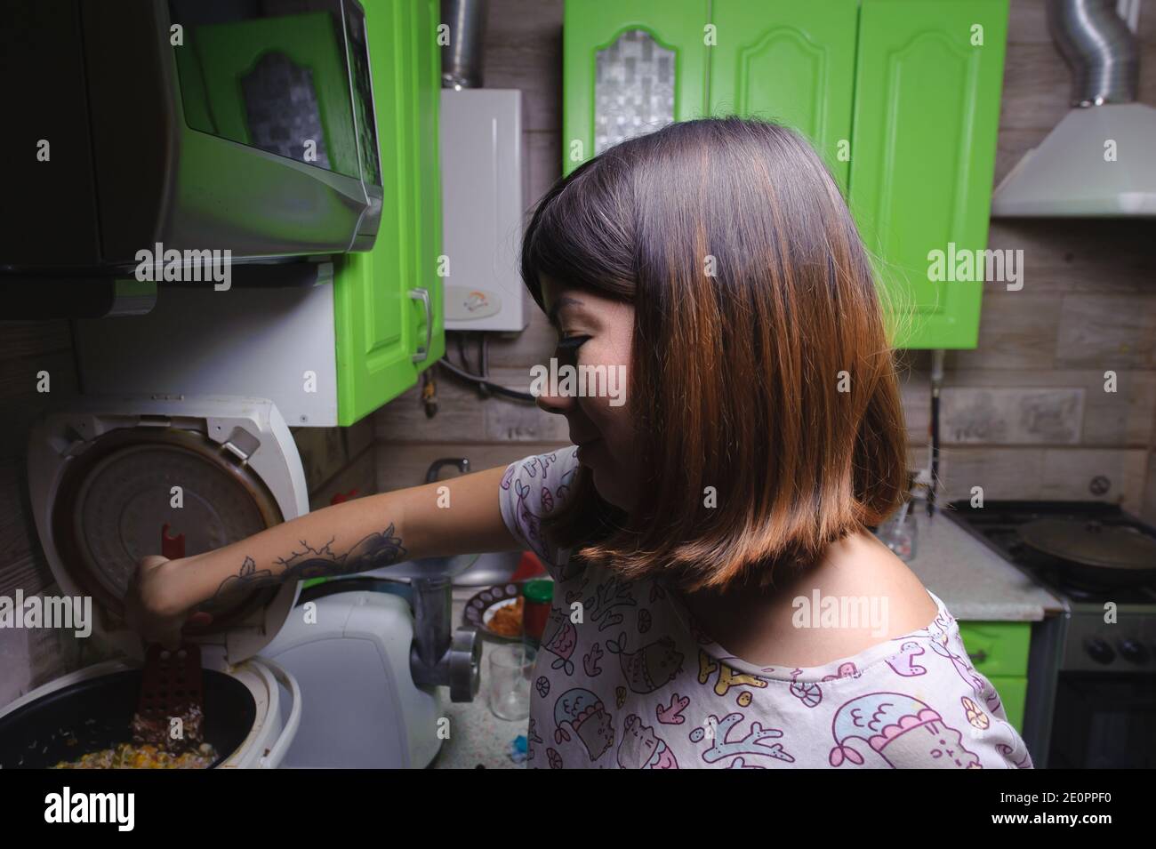 Lächelnde junge schwarzhaarige Frau, die Reis mit Gemüse mit einem Küchenschaber Spatel rührt, der für das Abendessen in einem Multikocker im k zubereitet wird Stockfoto