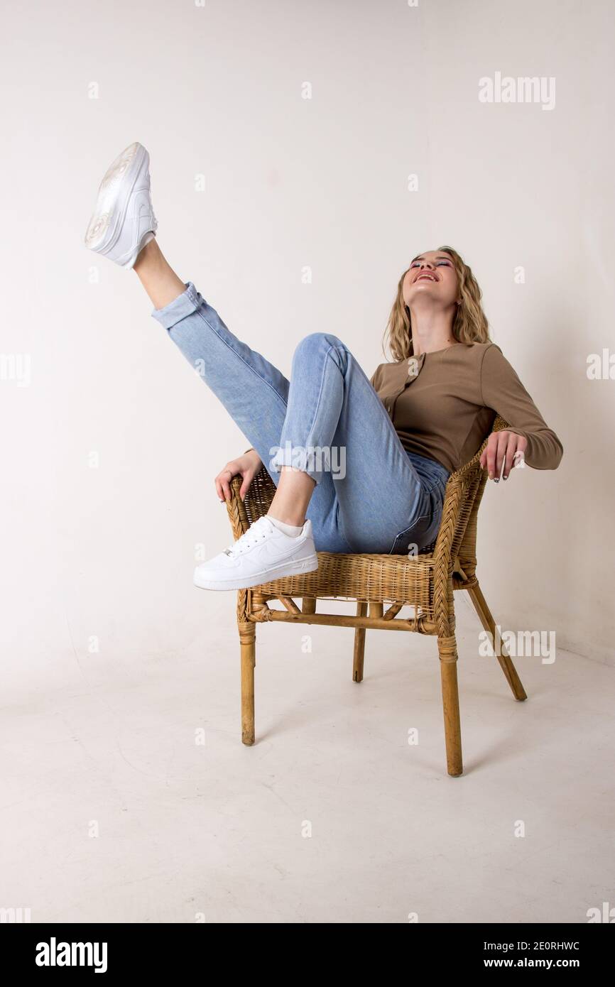 Porträt einer schönen jungen Frau in Jeans, Hemd und Turnschuhen.Sie legte ihre Füße auf den Weidenschaukelstuhl gegen die weiße Ziegelwand.sitzend Stockfoto