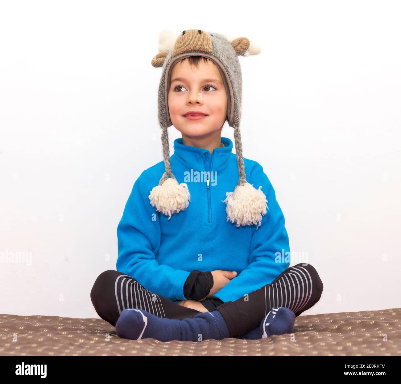 Glückliches Kind sitzt mit gekreuzten Beinen auf dem Bett zu Hause, in Winterkleidung und mit einem elchenförmigen Wollhut, der seinen Kopf warm hält. Stockfoto
