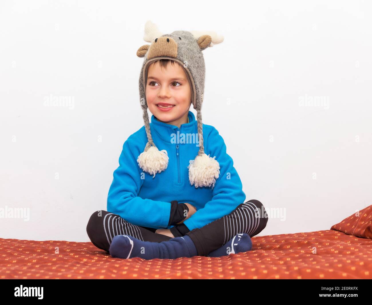 Glückliches Kind sitzt mit gekreuzten Beinen auf dem Bett zu Hause, in Winterkleidung und mit einem elchenförmigen Wollhut, der seinen Kopf warm hält. Stockfoto