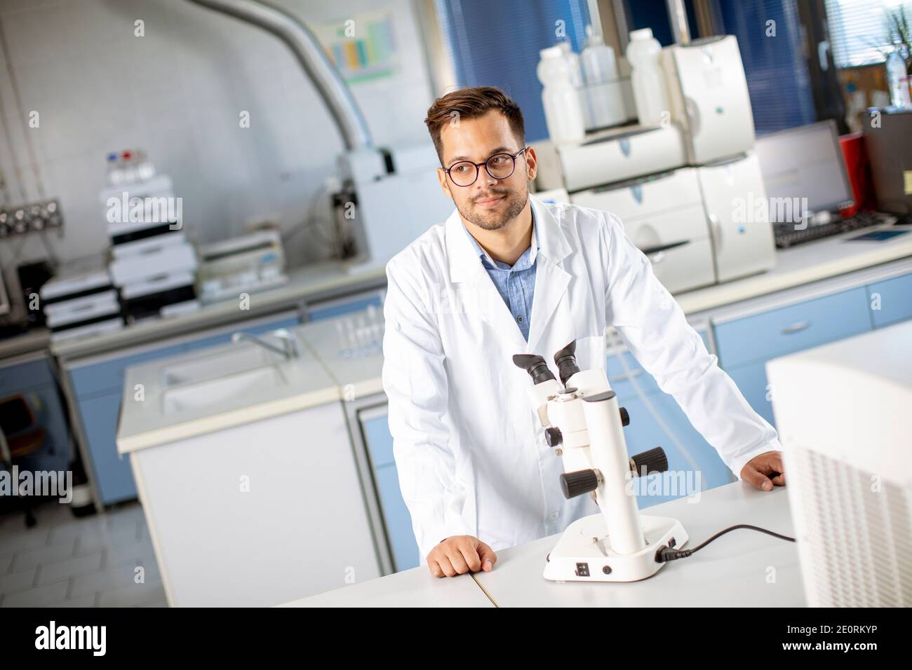 Hübscher junger Wissenschaftler im weißen Laborkittel, der mit binokular arbeitet Mikroskop im Labor der Materialwissenschaft Stockfoto