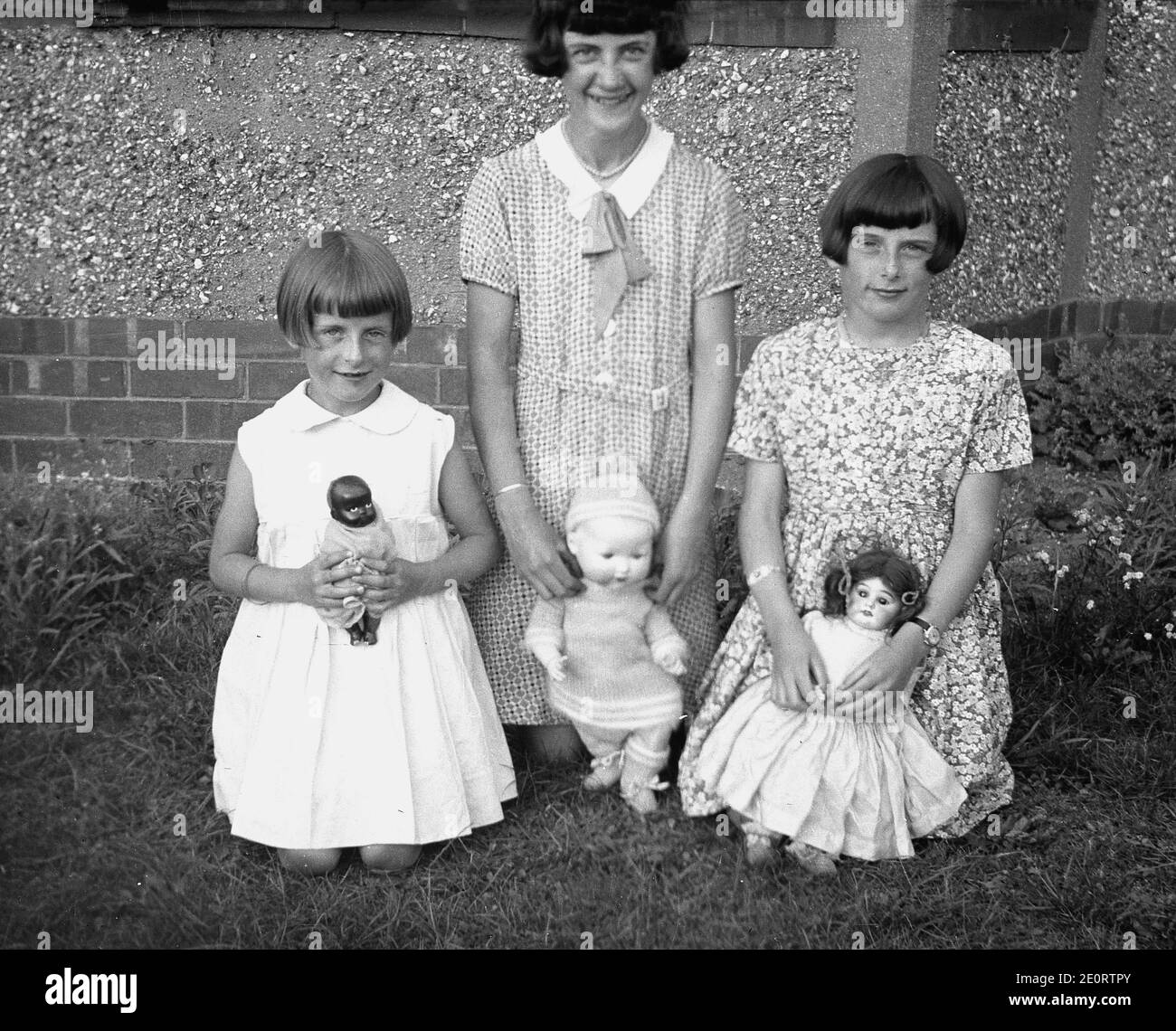 1940er Jahre, historisch, drei süße junge Mädchen, möglicherweise Schwestern, da sie den gleichen Stil der Frisur haben - bekannt als ein Puddingbecken Haarschnitt - kniend auf dem Gras vor einem Kieselstein-Dash Haus mit ihren wertvollen Spielzeugpuppen. Stockfoto