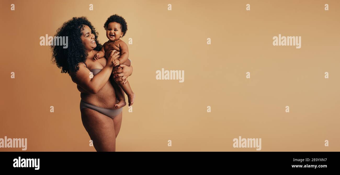 afroamerikanische Frau, die ihr Kind auf braunem Hintergrund in den Armen trägt. Fröhliche Mutter und Baby. Stockfoto