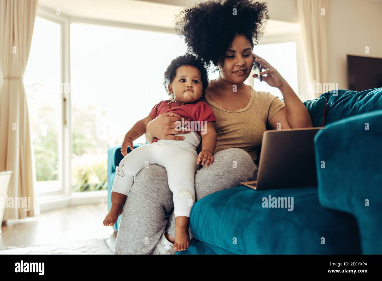 Afrikanische Mutter, die mit einem Baby auf dem Sofa sitzt und am Telefon telefoniert und mit einem Laptop arbeitet. Stockfoto