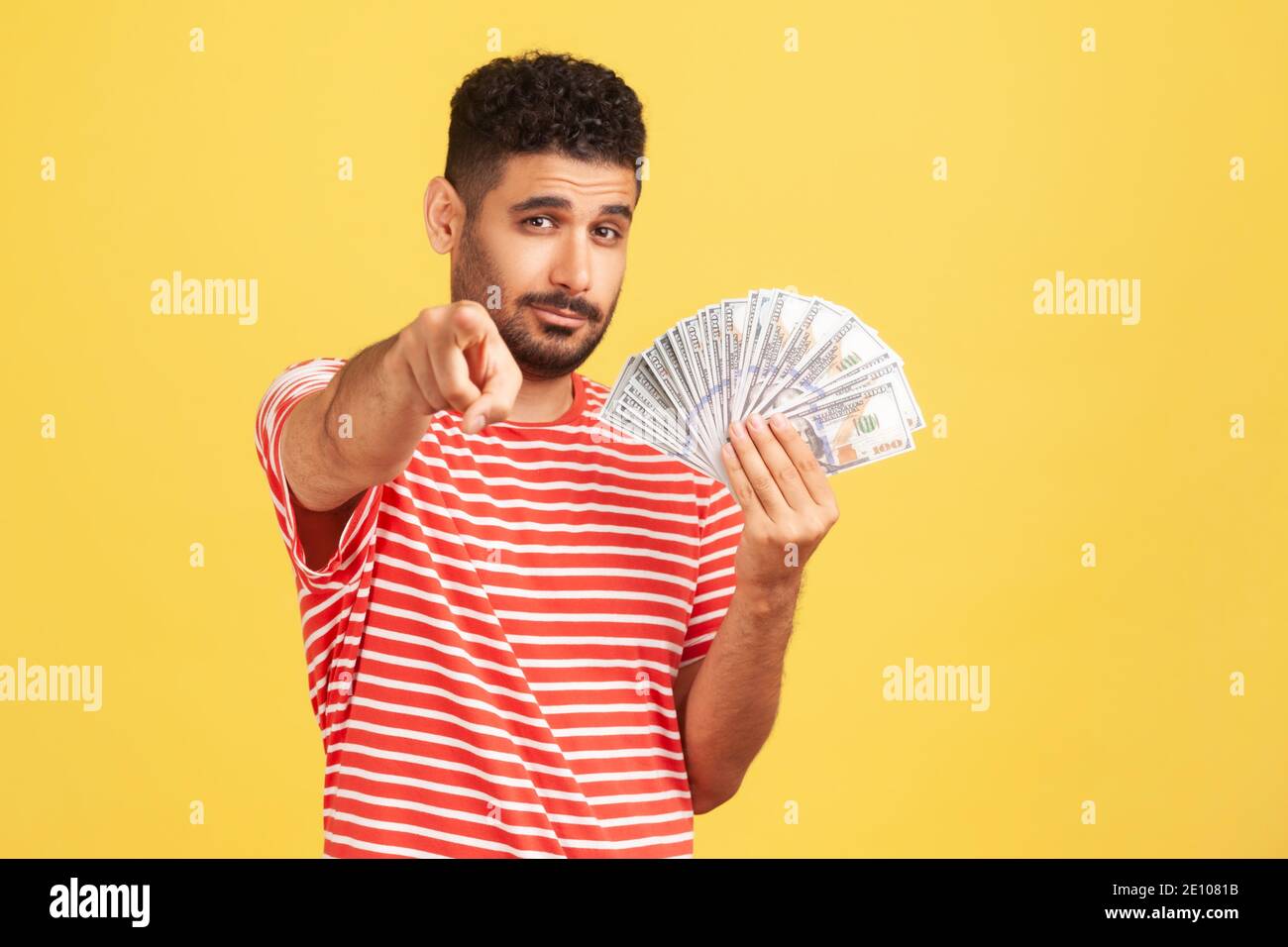 Selbstbewusster reicher Mann mit Bart in gestreiftem T-Shirt hält Fan von Dollar und zeigt Finger auf die Kamera mit ernster Ausdruck, einfacher Gewinn. Indoo Stockfoto