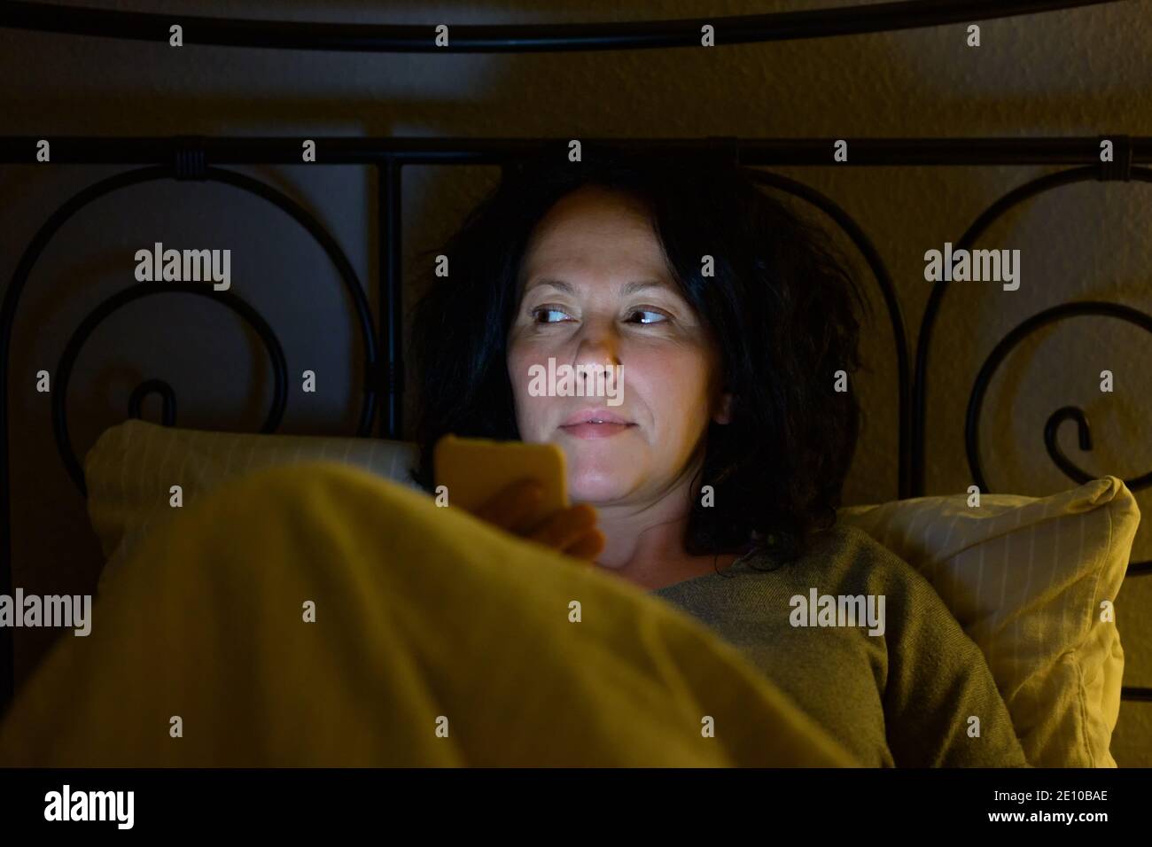Nachdenkliche hispanische Frau, die sich nachts im Bett entspannt, mit Fokus Zu ihrem Gesicht durch den Schein ihres Smartphones beleuchtet Als sie Seite anstarte Stockfoto