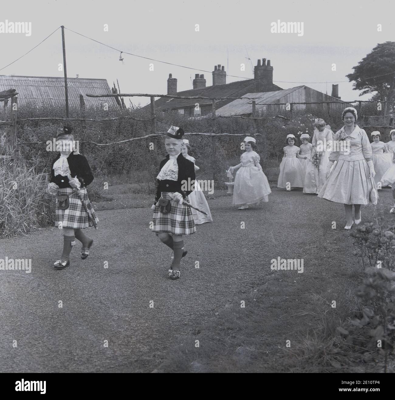 1950, historisch, zwei junge Jungen in Kilts führen die Rose Queen und ihre Gefolge von jungen Mädchen in ihren Kleidern durch einen lokalen Park, zu dem die Zeremonie stattfinden wird, Farnworth, Lancashire, England, Großbritannien. Der Tag der Rosenkönigin war ein besonderer Tag für die Kinder der Stadt, die an der Sonntagsschule der Kirche teilnahmen und mit der traditionellen "Sommersonnenwende" verbunden waren, die ihren Ursprung in den alten heidnischen Festen hatte. Es wurde manchmal zusammen mit dem Walking Day getan, wenn die Kinder versammelten sich und gingen durch die Straßen. Stockfoto