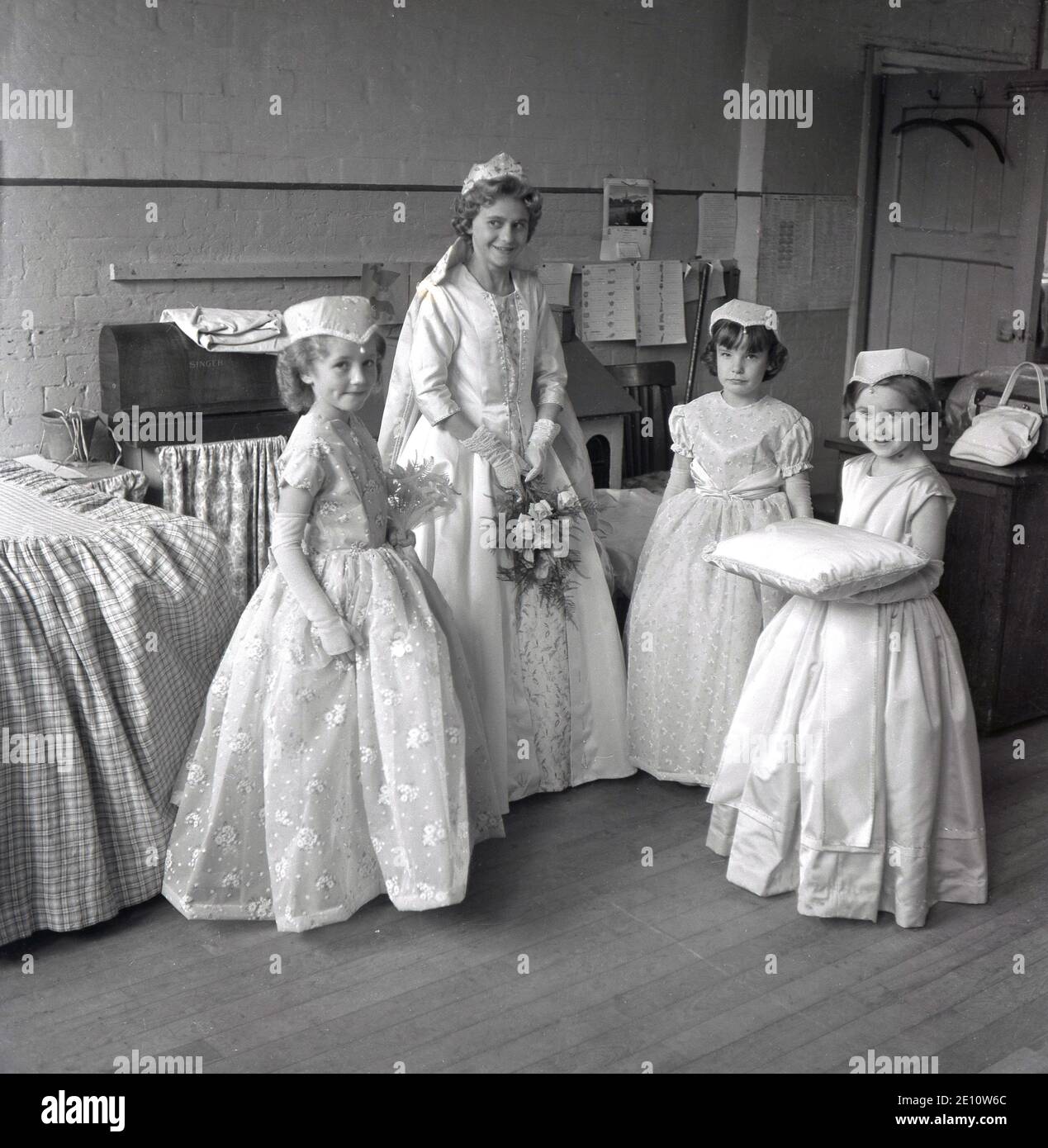 1950s, historisch, eine junge Rose Queen in ihrem Kleid mit ihrem kleinen weiblichen Gefolge gekleidet, eine hält das Kissen für ihre Krone, zusammen in der Dorfhalle nach der Zeremonie und Krönung, Farrnworth, Lancashire, England, Großbritannien. Die Krönung der Rosenkönigin war ein besonderer Tag für die Kinder der Stadt, die die Sonntagsschule der Kirche besuchten und mit der traditionellen "Sommersonnenwende" verbunden waren, die ihren Ursprung in den alten heidnischen Festen hatte. Es wurde manchmal zusammen mit dem Walking Day getan, wenn die Kinder versammelten sich und gingen durch die Straßen. Stockfoto