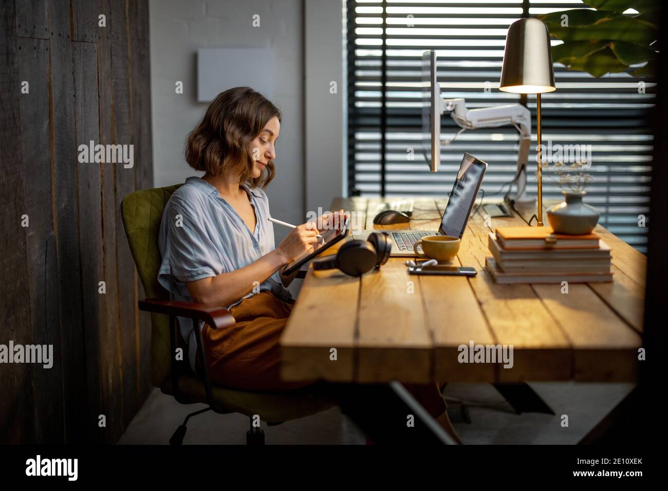 Junge Frau, die lässig gekleidet ist und kreative Arbeit hat, auf einem digitalen Tablet zeichnet und im gemütlichen und stilvollen Home Office sitzt Stockfoto