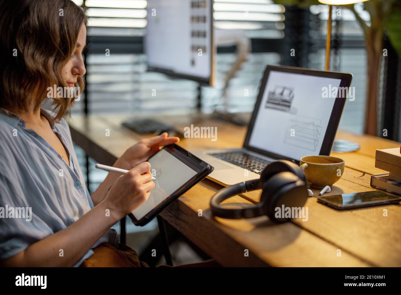 Junge Frau, die lässig gekleidet ist und kreative Arbeit hat, auf einem digitalen Tablet zeichnet und im gemütlichen und stilvollen Home Office sitzt Stockfoto