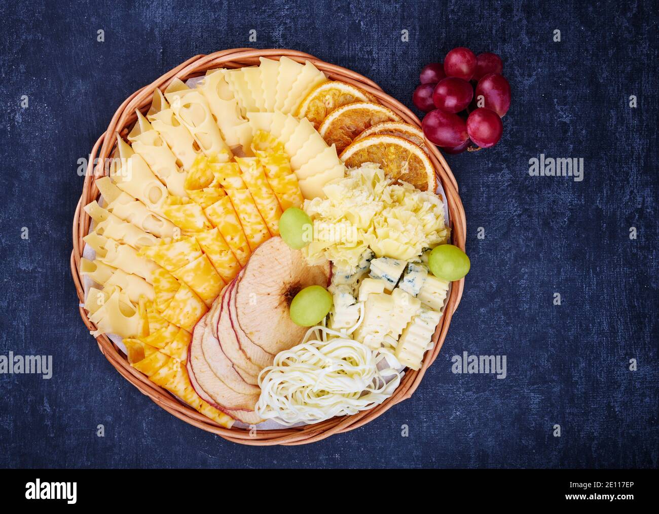 Weidenkorb mit einer köstlichen Auswahl an Käse, getrockneten Orangen- und Apfelscheiben auf dunklem Hintergrund Stockfoto