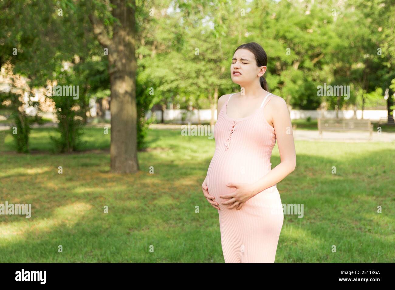 Eine schwangere junge Frau im Park und hält sich an den Magen, der schmerzt. Schwangerschaft und Mutterschaft. Rückenschmerzen während der Schwangerschaft. Stockfoto