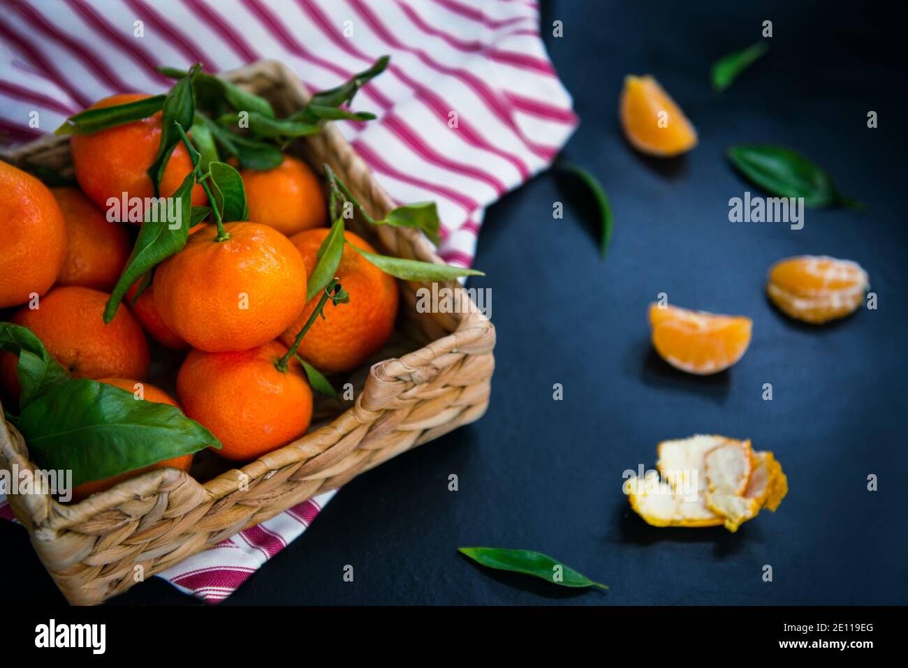 Frische reife Mandarinen, Clementine, Mandarine mit grünen Blättern in der Strohschale auf gestreifter Serviette und dunkelblauem Steinhintergrund. Lokale Zitrusfrüchte Stockfoto