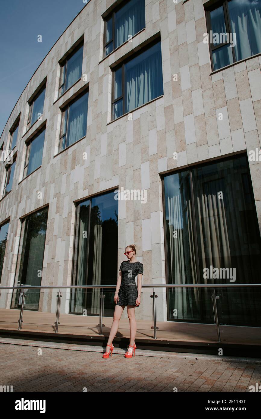 Das hübsche blonde Mädchen steht vor einem modernen Stadtgebäude; sie trägt ein stilvolles kurzes dunkles Kleid, eine rote Sonnenbrille und High Heels Stockfoto
